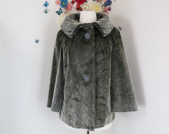 May include: A vintage olive green faux fur capelet with a large collar and three button closure. The capelet is displayed on a mannequin against a white wall decorated with colorful flowers and butterflies.