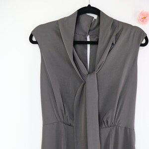 Puede incluir: Un vestido gris sin mangas con cuello drapeado y detalle de lazo. El vestido est&aacute; colgado de una percha negra sobre un fondo blanco. El vestido tiene cintura entallada y falda recta.