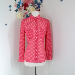 May include: A vibrant pink blouse with a white polka dot design. The long-sleeved shirt features a collar, button-down front, and two chest pockets. The garment is displayed on a mannequin.