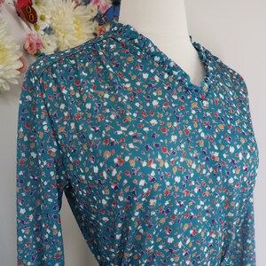 May include: A long-sleeved dress in a teal blue base color featuring a small-scale abstract floral print in white, red, and purple. The dress has a V-neckline with a ruffled collar detail and includes a matching fabric tie belt at the waist.