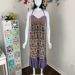 Peut inclure: Une robe à fleurs avec un col licou et un ourlet à volants à carreaux violets et blancs. La robe présente un corsage à motifs violets et roses et un bandeau assorti. La robe est présentée sur un mannequin.