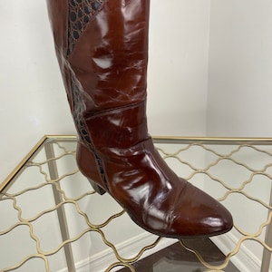 May include: A tall, brown leather boot with a pointed toe. The upper section features a textured, dark brown panel. The boot is knee-high and has a low heel. The leather has a glossy finish.