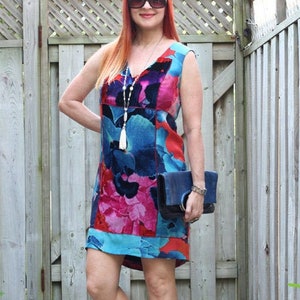 Pode incluir: Um vestido sem mangas, na altura do joelho, com um padr&atilde;o floral vibrante em tons de azul, rosa e vermelho. O vestido &eacute; combinado com uma clutch azul escuro e saltos magenta. Um colar comprido com uma borla branca tamb&eacute;m &eacute; vis&iacute;vel.