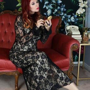 May include: A woman in a black lace dress sits on a red velvet sofa in front of a floral wallpaper. The dress has long sleeves and a floral pattern. She is holding a small, gold-colored compact mirror.