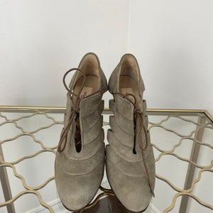 May include: A pair of taupe-colored suede heeled shoes with a lace-up design. The shoes have a unique cut-out pattern and are displayed on a gold-colored metal table.