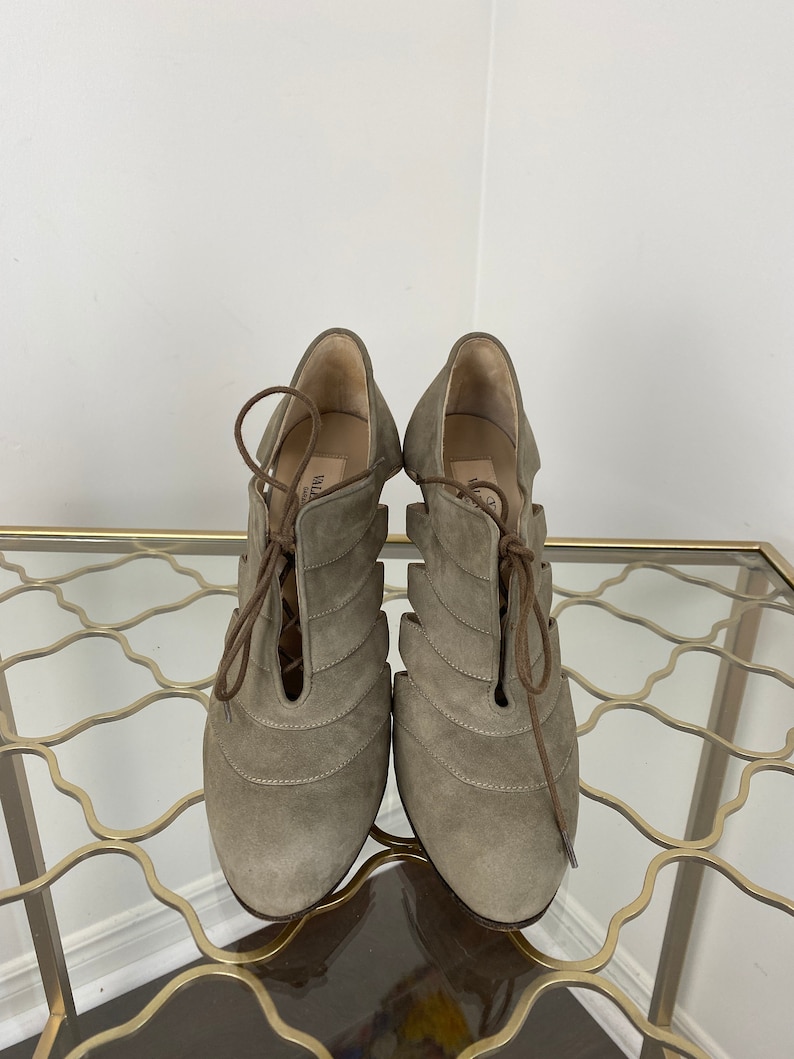 May include: A pair of taupe-colored suede heeled shoes with a lace-up design. The shoes have a unique cut-out pattern and are displayed on a gold-colored metal table.