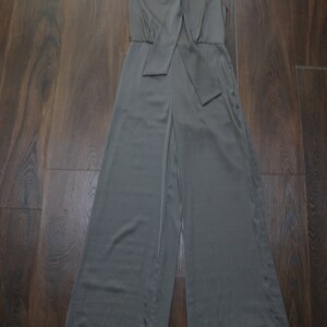 Puede incluir: Mono verde oliva sin mangas con perneras anchas, colgado de una percha negra. El mono presenta un cuello y un detalle de lazo en la cintura. La tela tiene un ligero brillo.