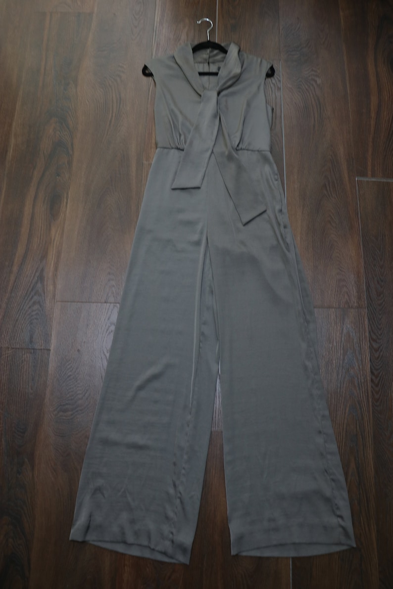 Puede incluir: Mono verde oliva sin mangas con perneras anchas, colgado de una percha negra. El mono presenta un cuello y un detalle de lazo en la cintura. La tela tiene un ligero brillo.