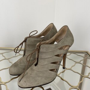 May include: A pair of taupe-colored suede high-heeled shoes with cut-out detailing and lace-up closure. The heels are approximately 4 inches tall. The shoes are displayed on a glass surface.