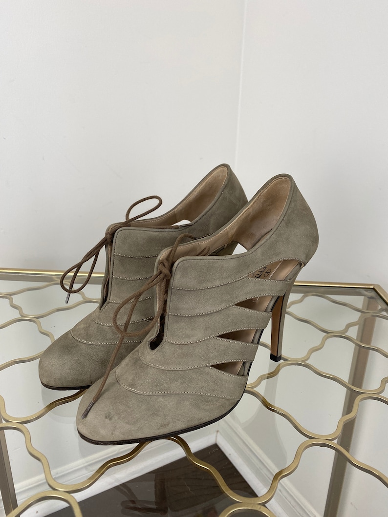 May include: A pair of taupe-colored suede high-heeled shoes with cut-out detailing and lace-up closure. The heels are approximately 4 inches tall. The shoes are displayed on a glass surface.