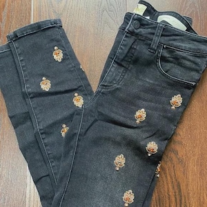 May include: Dark grey denim jeans featuring embroidered floral patterns in gold and orange. The jeans have a classic five-pocket design and a button and zip closure.