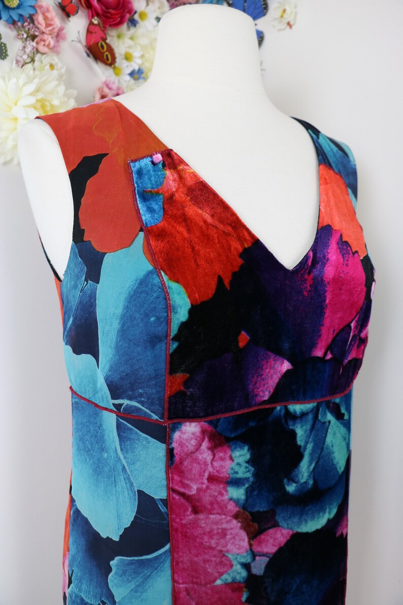 Pode incluir: Vestido sem mangas com decote em V, apresentando um vibrante padr&atilde;o floral em tons de azul, vermelho e rosa. O vestido &eacute; exibido em um manequim, mostrando seu design e paleta de cores. O tecido parece ter uma textura aveludada.