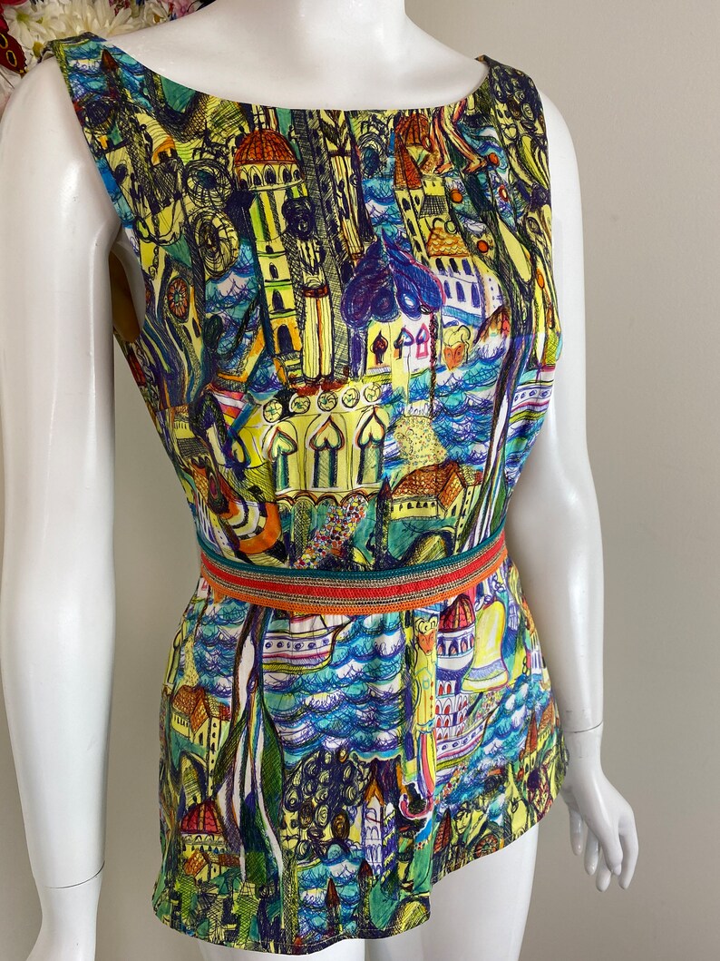 May include: Sleeveless top with a vibrant cityscape print. The design features buildings, water, and boats in shades of yellow, blue, and green. A decorative belt with orange and green accents cinches the waist.
