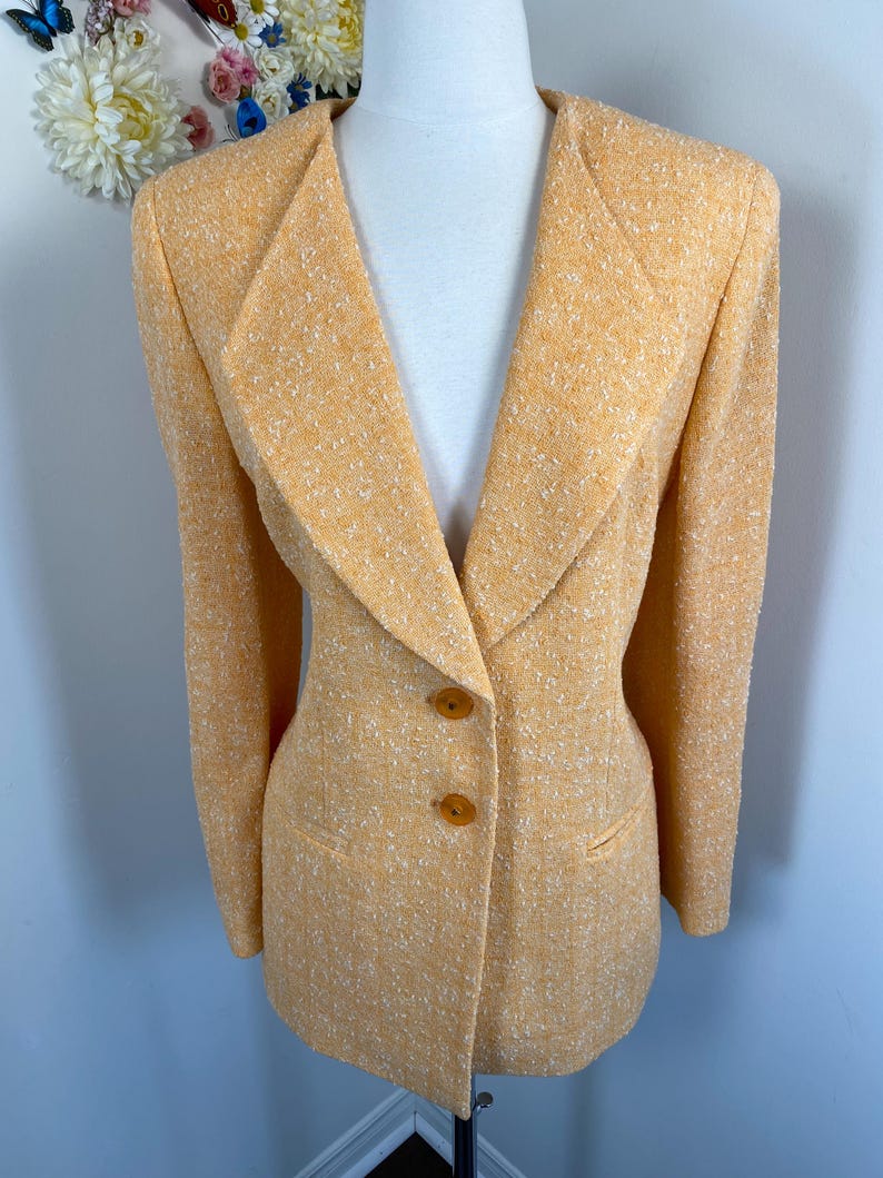 May include: A light orange blazer with a textured fabric. The blazer has a notched collar, two brown buttons, and two front pockets. The jacket is displayed on a mannequin.