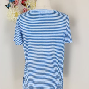 May include: A short-sleeved, blue and white striped shirt. The shirt is displayed on a mannequin. The background features a floral arrangement with butterflies. The shirt's design is a classic horizontal stripe pattern.