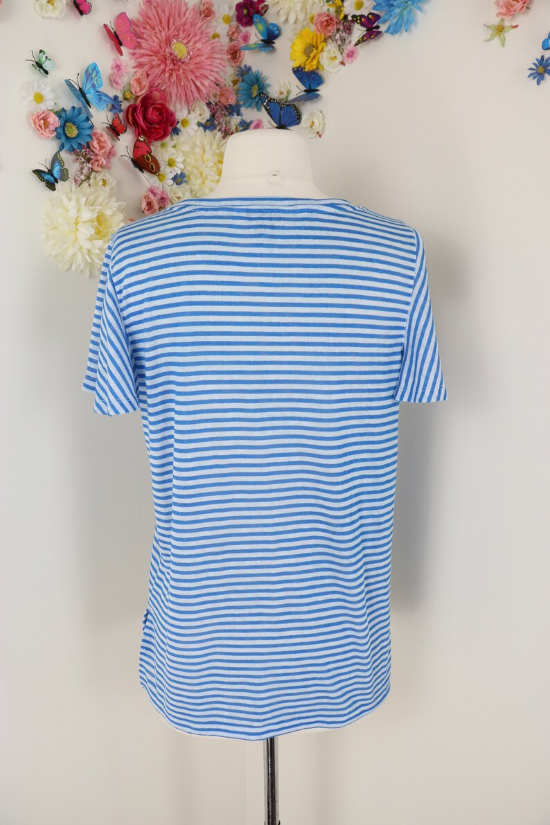 May include: A short-sleeved, blue and white striped shirt. The shirt is displayed on a mannequin. The background features a floral arrangement with butterflies. The shirt's design is a classic horizontal stripe pattern.