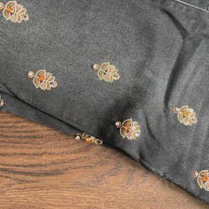May include: Close-up of a dark grey fabric with embroidered floral patterns. The embroidery features gold, orange, and clear bead accents. The fabric is folded, revealing a glimpse of a wooden surface.