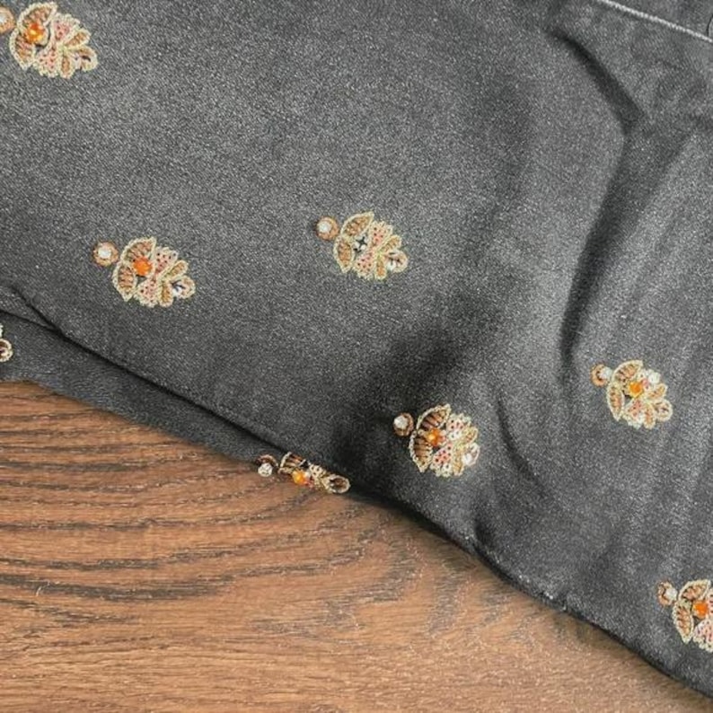 May include: Close-up of a dark grey fabric with embroidered floral patterns. The embroidery features gold, orange, and clear bead accents. The fabric is folded, revealing a glimpse of a wooden surface.