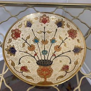 May include: A decorative round plate with a floral design. The plate features a cream-colored background with gold accents and a central vase filled with colorful flowers in red, blue, and gold. The plate is displayed on a gold-colored metal stand.