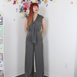 Puede incluir: Mono gris con detalle de lazo en el escote. El mono sin mangas tiene perneras anchas y un corte relajado. El fondo presenta una pared decorada con flores y mariposas de colores.