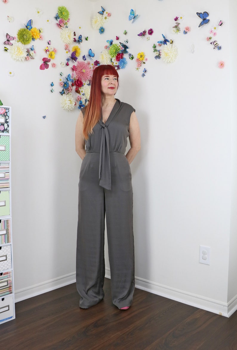 Puede incluir: Mono gris con detalle de lazo en el escote. El mono sin mangas tiene perneras anchas y un corte relajado. El fondo presenta una pared decorada con flores y mariposas de colores.