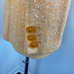 May include: Close-up of a yellow tweed blazer with three amber-colored buttons. The blazer has a textured weave with white flecks throughout. The garment is displayed on a silver mannequin.