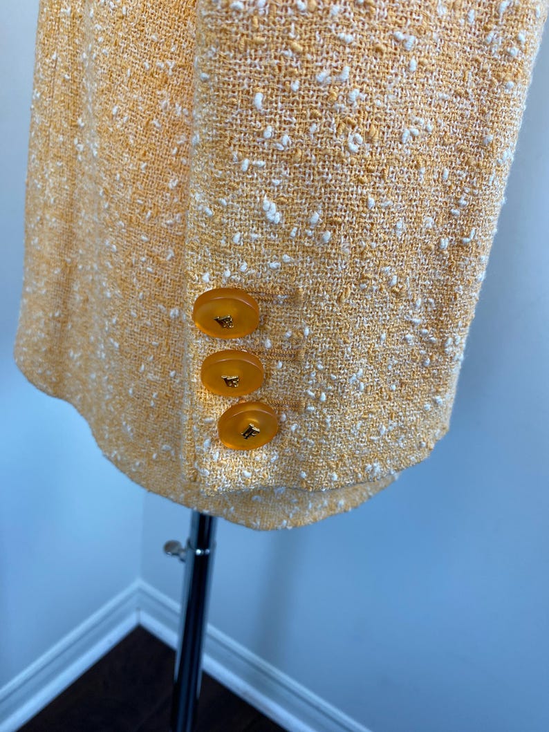 May include: Close-up of a yellow tweed blazer with three amber-colored buttons. The blazer has a textured weave with white flecks throughout. The garment is displayed on a silver mannequin.