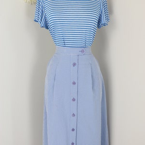 May include: A two-piece outfit on a mannequin. The top is a light blue and white striped t-shirt. The bottom is a light blue gingham skirt with buttons down the front. The outfit is displayed against a white background.