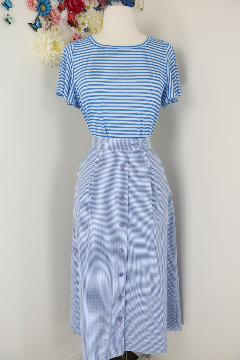 May include: A two-piece outfit on a mannequin. The top is a light blue and white striped t-shirt. The bottom is a light blue gingham skirt with buttons down the front. The outfit is displayed against a white background.