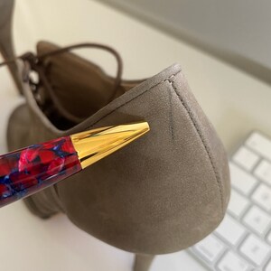 May include: Close-up of a taupe-colored suede shoe with a small heel. The shoe has a lace-up design. A pen with a red and blue marbled design and a gold-colored tip is in the foreground.