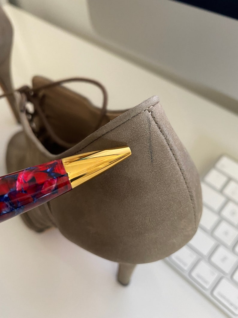 May include: Close-up of a taupe-colored suede shoe with a small heel. The shoe has a lace-up design. A pen with a red and blue marbled design and a gold-colored tip is in the foreground.