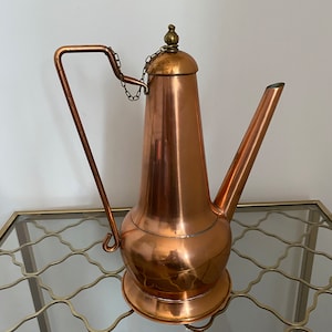 May include: A polished copper pitcher with a tall, tapered body, a wide rounded base, and a long, angled spout. It features a thin, angular metal handle connected to the lid by a small chain, and a decorative brass finial sits atop the lid.