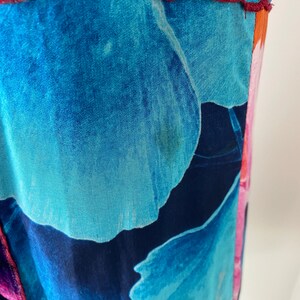 Pode incluir: Close-up de uma pe&ccedil;a de roupa com um padr&atilde;o floral vibrante. O tecido apresenta grandes p&eacute;talas de flores azuis e turquesas estilizadas contra um fundo azul marinho profundo. O design inclui detalhes em rosa e laranja, com uma costura bord&ocirc;.