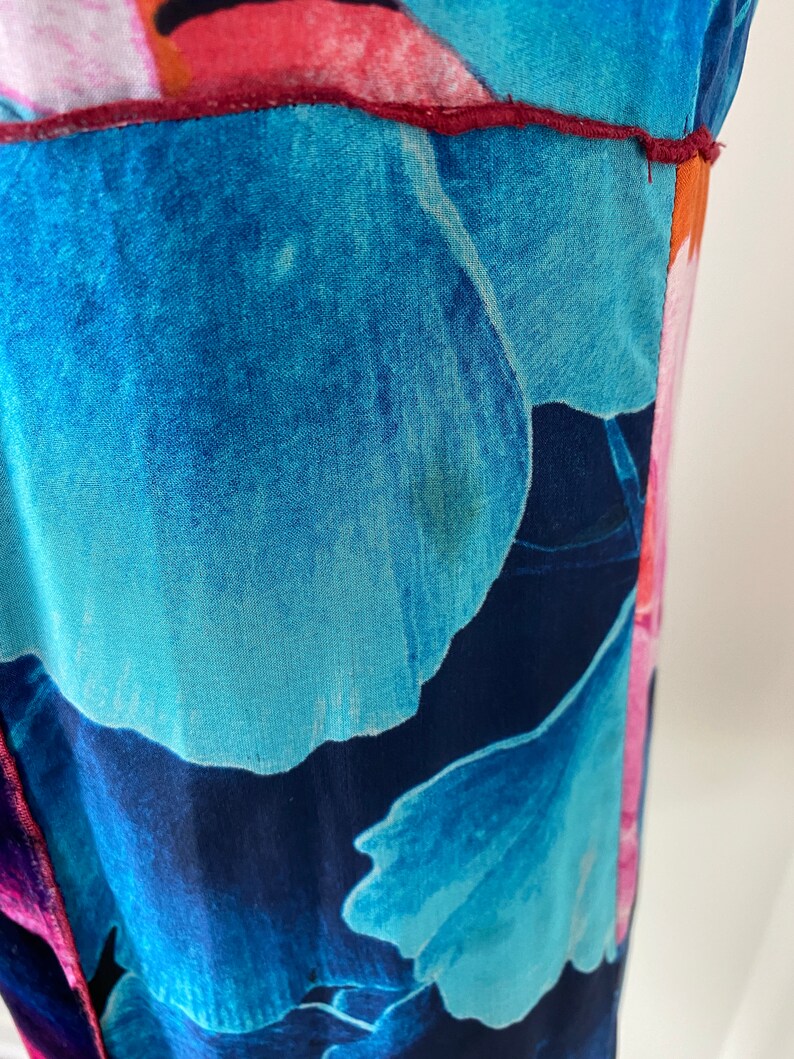Pode incluir: Close-up de uma pe&ccedil;a de roupa com um padr&atilde;o floral vibrante. O tecido apresenta grandes p&eacute;talas de flores azuis e turquesas estilizadas contra um fundo azul marinho profundo. O design inclui detalhes em rosa e laranja, com uma costura bord&ocirc;.