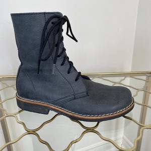 Peut inclure: Une botte à lacets en cuir bleu foncé avec un bout arrondi, une hauteur mi-mollet et des lacets noirs. La botte possède une semelle noire épaisse avec une trépointe claire visible et des coutures blanches le long de la base de la chaussure.