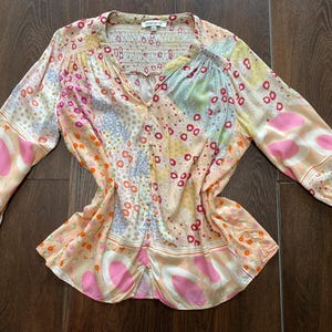 May include: A patterned blouse with long sleeves and a V-neck. The design incorporates pink, orange, yellow, and blue patches. The sleeves have elasticated cuffs. The fabric appears to be lightweight, suitable for warmer weather.