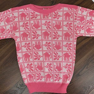 Peut inclure: Un pull rose à manches courtes avec un motif répété d'éléphants et de motifs floraux en rose clair. Le pull a un col, des poignets et un ourlet côtelés. Le motif est disposé en grille.