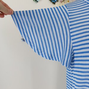May include: Close-up of a blue and white striped t-shirt sleeve. The sleeve is made of a lightweight fabric and has a flared design. The stripes are vertical and evenly spaced. The shirt is a light blue and white color.
