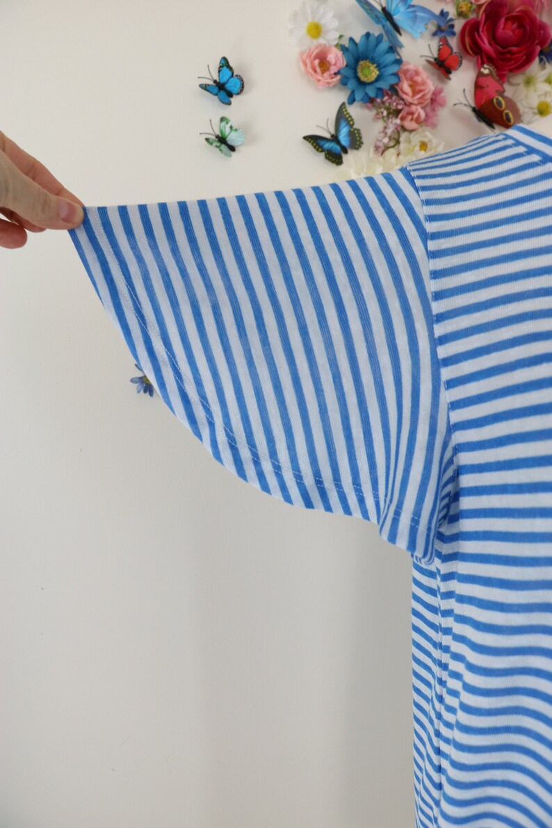 May include: Close-up of a blue and white striped t-shirt sleeve. The sleeve is made of a lightweight fabric and has a flared design. The stripes are vertical and evenly spaced. The shirt is a light blue and white color.