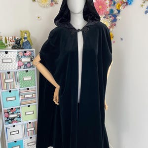 May include: A black velvet hooded cloak with a clasp closure. The cloak is displayed on a mannequin. The cloak has a hood and a flowing design. The background features a floral arrangement and a decorative storage unit.