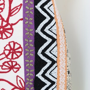 May include: Close-up of a textile with a variety of patterns and textures. Features include red floral embroidery on white fabric, a purple stripe with green accents, a black beaded zigzag design, and an orange stripe with a white pattern.