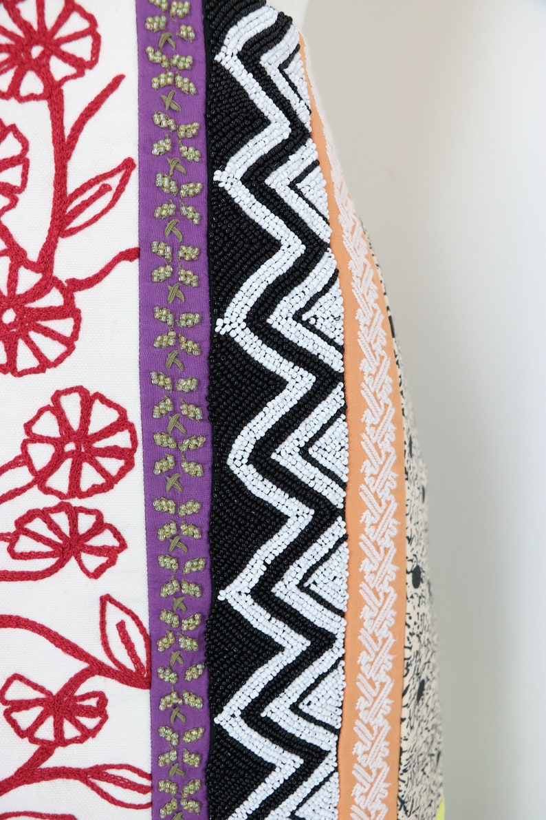 May include: Close-up of a textile with a variety of patterns and textures. Features include red floral embroidery on white fabric, a purple stripe with green accents, a black beaded zigzag design, and an orange stripe with a white pattern.