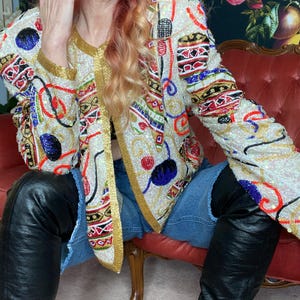 May include: A long-sleeve jacket featuring an off-white base covered in intricate, multi-colored beadwork and sequins. The design includes gold trim along the front opening and cuffs, with abstract patterns in red, blue, black, and green.