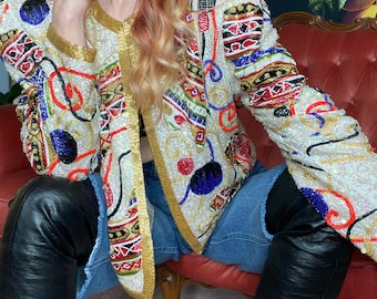 May include: A long-sleeve jacket featuring an off-white base covered in intricate, multi-colored beadwork and sequins. The design includes gold trim along the front opening and cuffs, with abstract patterns in red, blue, black, and green.