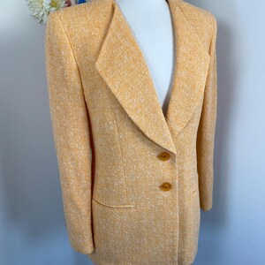 May include: A light orange blazer with a textured weave. The blazer features a notched lapel, two brown buttons, and a single pocket. The jacket is displayed on a mannequin.