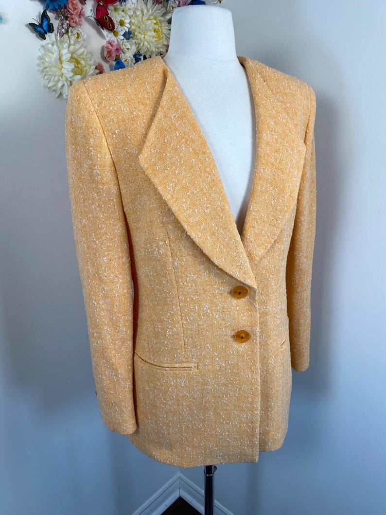 May include: A light orange blazer with a textured weave. The blazer features a notched lapel, two brown buttons, and a single pocket. The jacket is displayed on a mannequin.