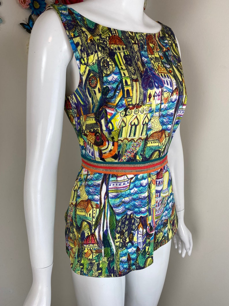 May include: Sleeveless top with a colourful cityscape print. The top features a boat neckline and a fitted waist with a decorative band. The print includes buildings, boats, and water in shades of blue, yellow, green, and purple.