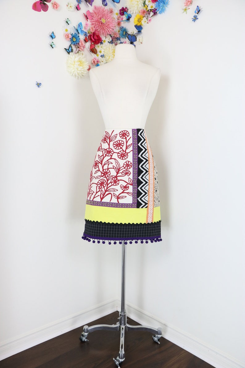 May include: A colorful, patterned skirt displayed on a mannequin. The skirt features a floral design in red, a yellow scalloped hem, and a black and white checkered bottom with purple pom-poms. The skirt is on a chrome stand.