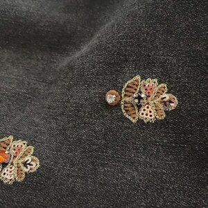 May include: Close-up of dark grey fabric with two embroidered floral designs. The flowers have brown, pink, and black petals with gold thread outlining the design. A small, round, sparkling gem is near each flower.
