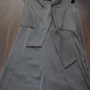 Puede incluir: Mono sin mangas verde oliva con cuello drapeado y detalle de lazo a juego. La prenda cuelga de una percha negra. La tela tiene un acabado liso, satinado. El fondo es un suelo de madera.